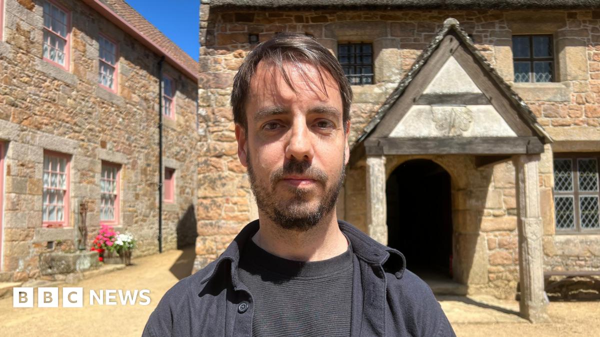 Ben Spink stands centered in a sunlit stone courtyard, facing the camera, wearing a dark shirt and jacket. Behind him are rustic buildings made of warm-toned stone, including one with pink-trimmed windows on the left and another with a thatched roof and a small wooden porch on the right. Bright blue sky and a pot of red and white flowers add colour to the scene.