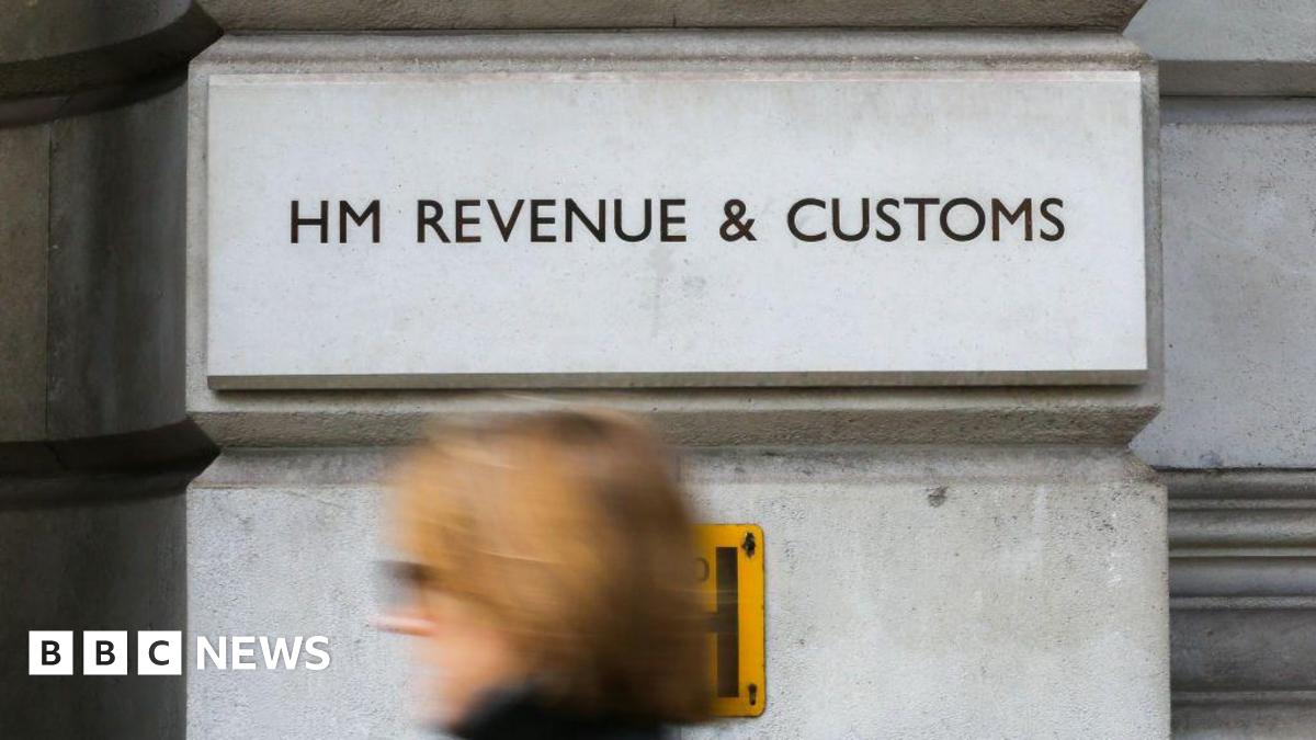 The HM Revenue & Customs logo on a pale brick. A woman walks past in the foreground.