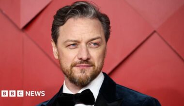 James McAvoy wearing a suit and a bow tie. There is a red background behind him.
