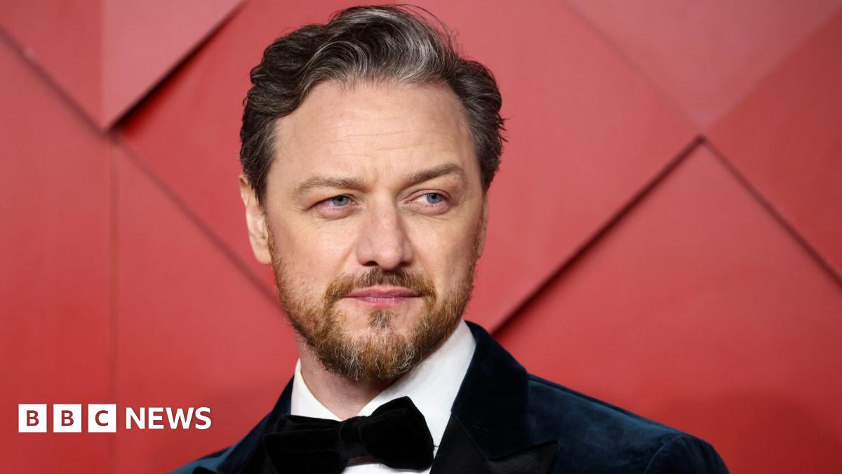 James McAvoy wearing a suit and a bow tie. There is a red background behind him.