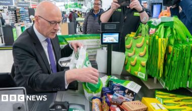 John Swinney is sitting at an Asda checkout where he is wearing a dark suit, white shirt and purple tie. He is bagging a pile of shopping as photographers and camera people capture the image.