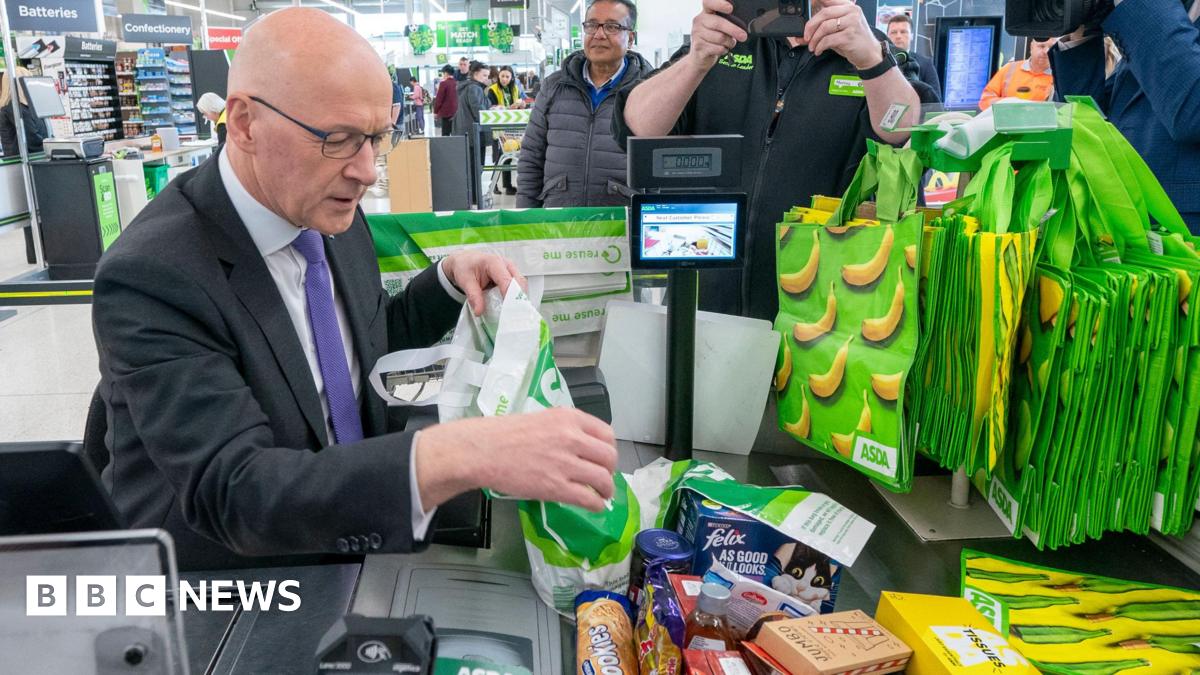 John Swinney is sitting at an Asda checkout where he is wearing a dark suit, white shirt and purple tie. He is bagging a pile of shopping as photographers and camera people capture the image.