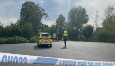 A police officer next to a police car in a car park with police cordon tape across the image