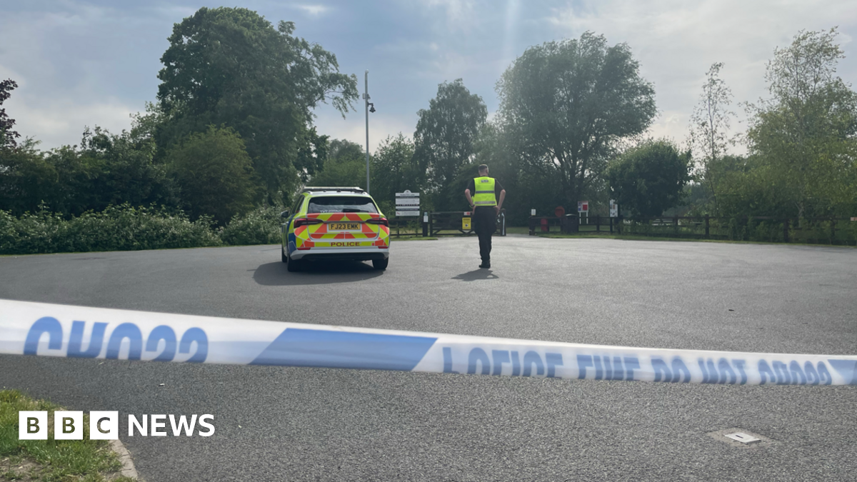 A police officer next to a police car in a car park with police cordon tape across the image