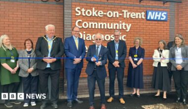 A man is cutting a blue ribbon with a line of people around him. Behind him is the wall of a building and a white sign which can be partly read and says "Stoke on Trent NHS Community Diagnostic".
