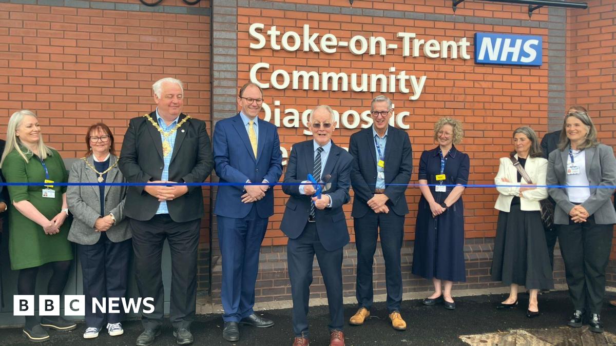 A man is cutting a blue ribbon with a line of people around him. Behind him is the wall of a building and a white sign which can be partly read and says "Stoke on Trent NHS Community Diagnostic".