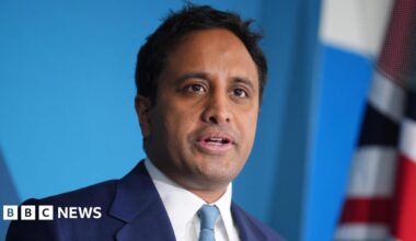 Zia Yusuf is stood in front of a blue background and a Union flag can be partially seen. He is wearing a dark blue suit, white shirt and a turquoise-coloured tie. He has short black hair.