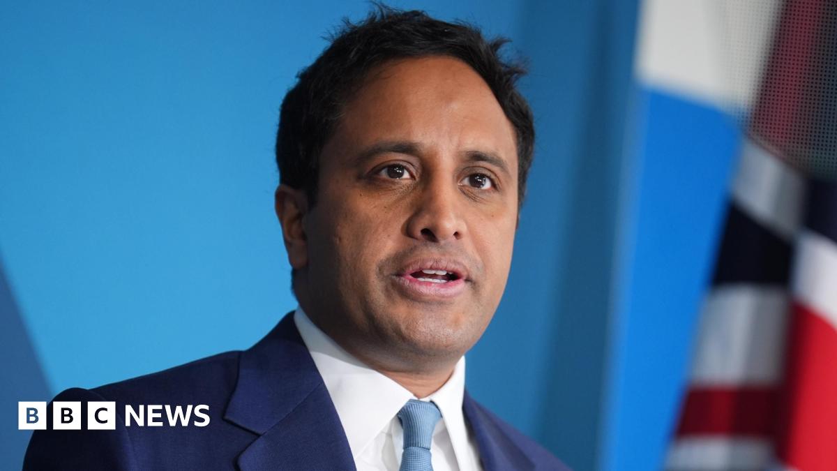 Zia Yusuf is stood in front of a blue background and a Union flag can be partially seen. He is wearing a dark blue suit, white shirt and a turquoise-coloured tie. He has short black hair.