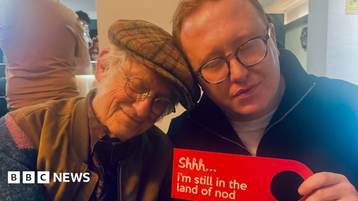 Noddy Holder in a tweed flat cap, glasses and a jacket has his eyes closed as he leans his head towards Tom Seals who also has his eyes closed and is wearing a black quarter zip jumper. He is holding a red sign that reads: Shhh... I'm still in the land of nod