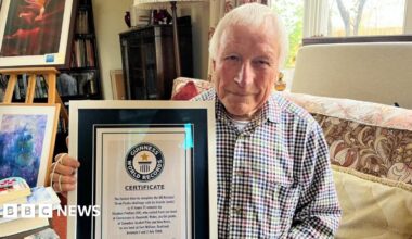 Wearing a checked shirt and with short white hair, Steve Poulton looks at the camera while holding the Guinness World Record certificate inside a frame. It reads "the fastest time to complete the UK national Three Peaks challenge by bicycle (male)".