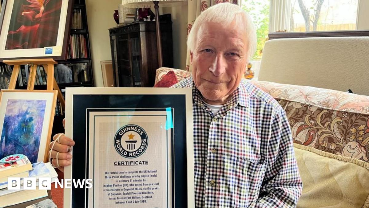 Wearing a checked shirt and with short white hair, Steve Poulton looks at the camera while holding the Guinness World Record certificate inside a frame. It reads "the fastest time to complete the UK national Three Peaks challenge by bicycle (male)".
