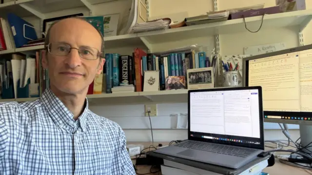 Richard Allan wears a blue striped shirt and glasses while sitting in his office