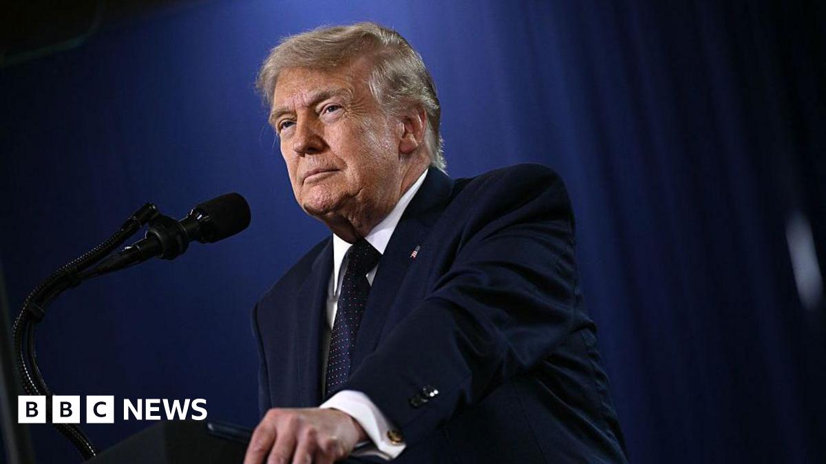 President Donald Trump wears a navy suit and tie and looks off into the distance with a microphone in front of him