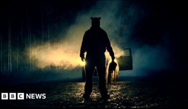 A silhouette of a figure in front of car headlights, at night. He is holding something ominous in his hand.