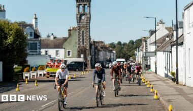 A group of cyclists pedal through the heart of the south of Scotland town of Gatehouse with houses down both sides of the road and traffic cones and a tower with a clock on it in the background.