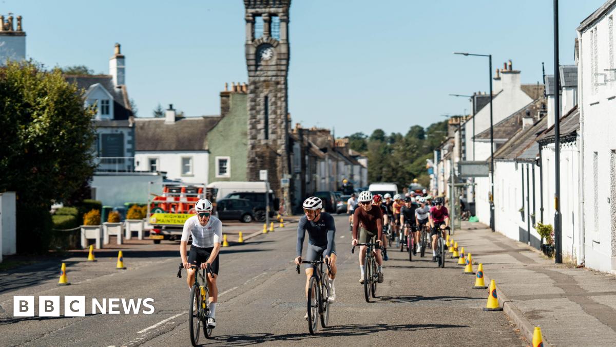 A group of cyclists pedal through the heart of the south of Scotland town of Gatehouse with houses down both sides of the road and traffic cones and a tower with a clock on it in the background.