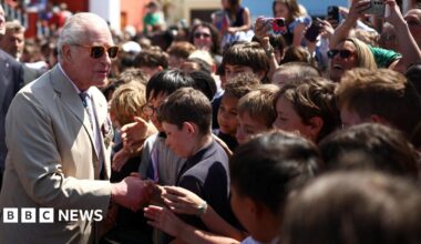 King Charles III wearing a beige suit with military medals, walks past a long row of Royal Bermuda Regiment troops