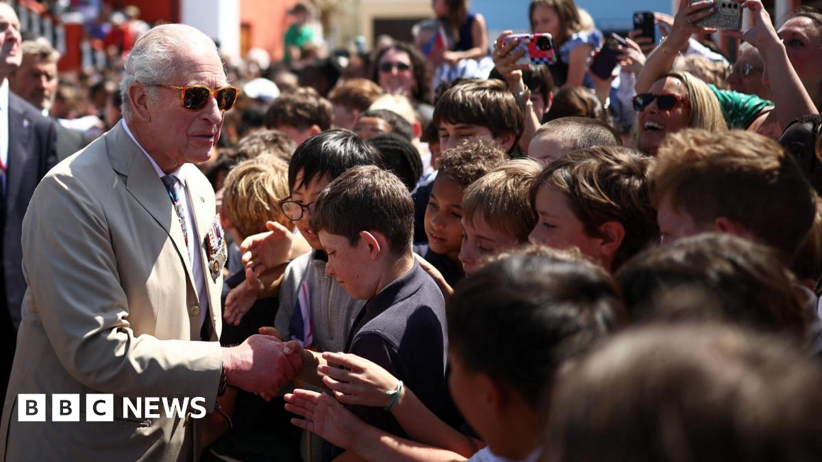 King Charles III wearing a beige suit with military medals, walks past a long row of Royal Bermuda Regiment troops