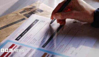 A woman in a wheelchair is sat in front of a laptop and a microphone, looking to her left. She has blonde hair, glasses and is wearing a bright red jumper.