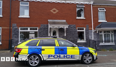A police car is parked outside a red and grey brick terrace house. Smashed windows are visible.