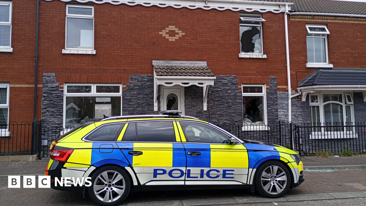 A police car is parked outside a red and grey brick terrace house. Smashed windows are visible.