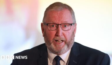 Jon Burrows is wearing a white shirt with navy blazer and tie. The background is blurry.