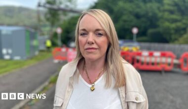 Ellen Forson, standing on a residential street in front of red safety barriers that block off the road, with trees and a hillside in the background. She is wearing a light-coloured jacket over a white top and a chain necklace, and is positioned in the foreground with the background softly out of focus.