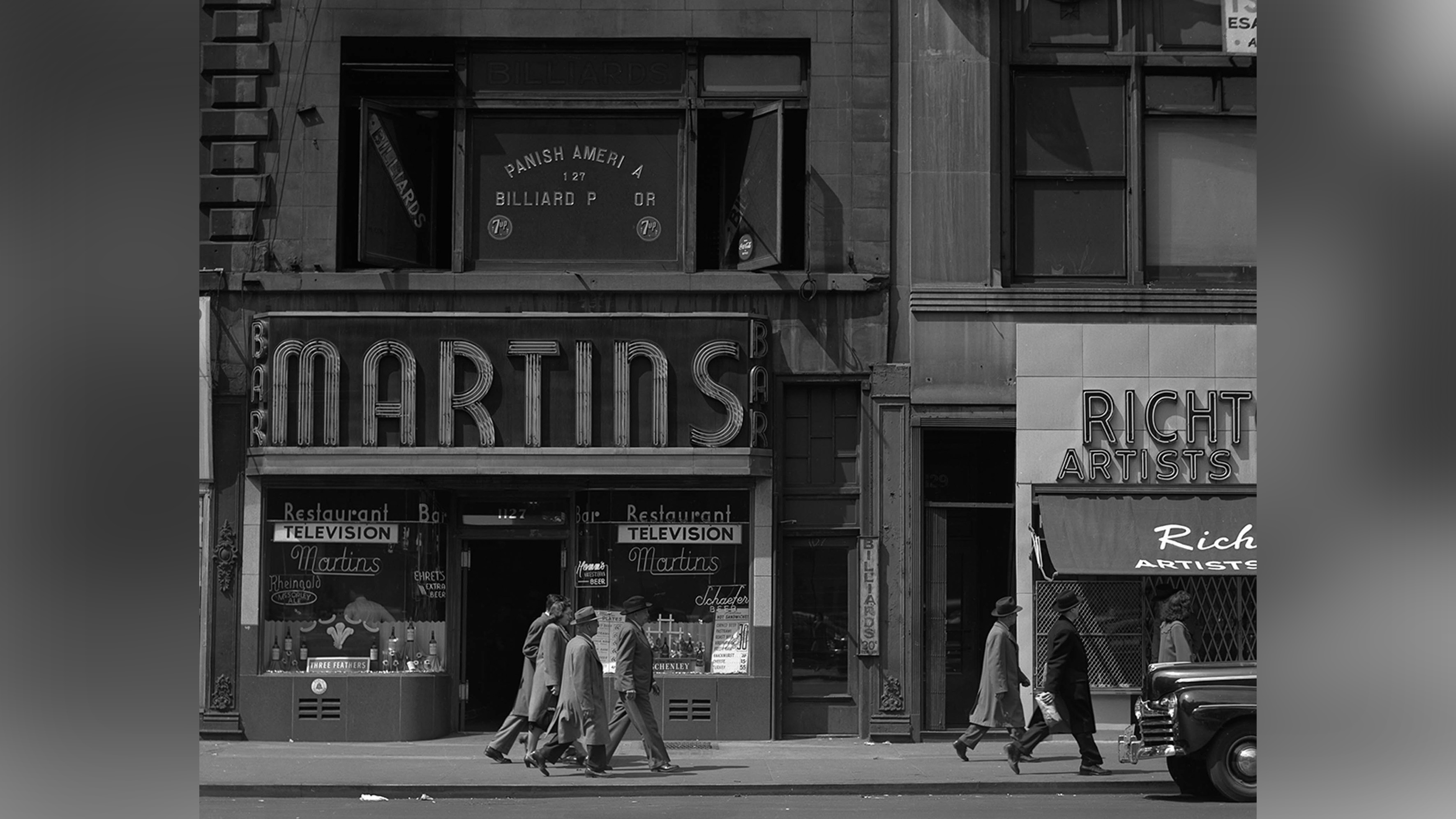 Group of men walk along pavement in front of shop.