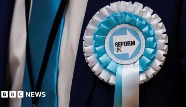 Close-up photo of a blue and white Reform UK rosette worn by someone wearing a dark suit.