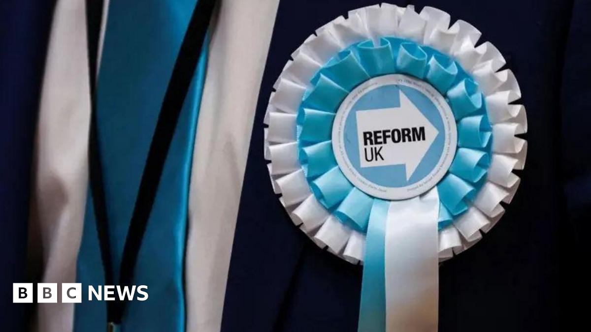 Close-up photo of a blue and white Reform UK rosette worn by someone wearing a dark suit.