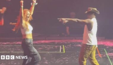 A woman with long red hair, a white tank top and black cargo trousers stands on a black stage stage with her arms in the air. To the right singer Ne-Yo, wearing a white vest top, mustard coloured trousers and a brown cowboy-style hat points towards her with his right hand.