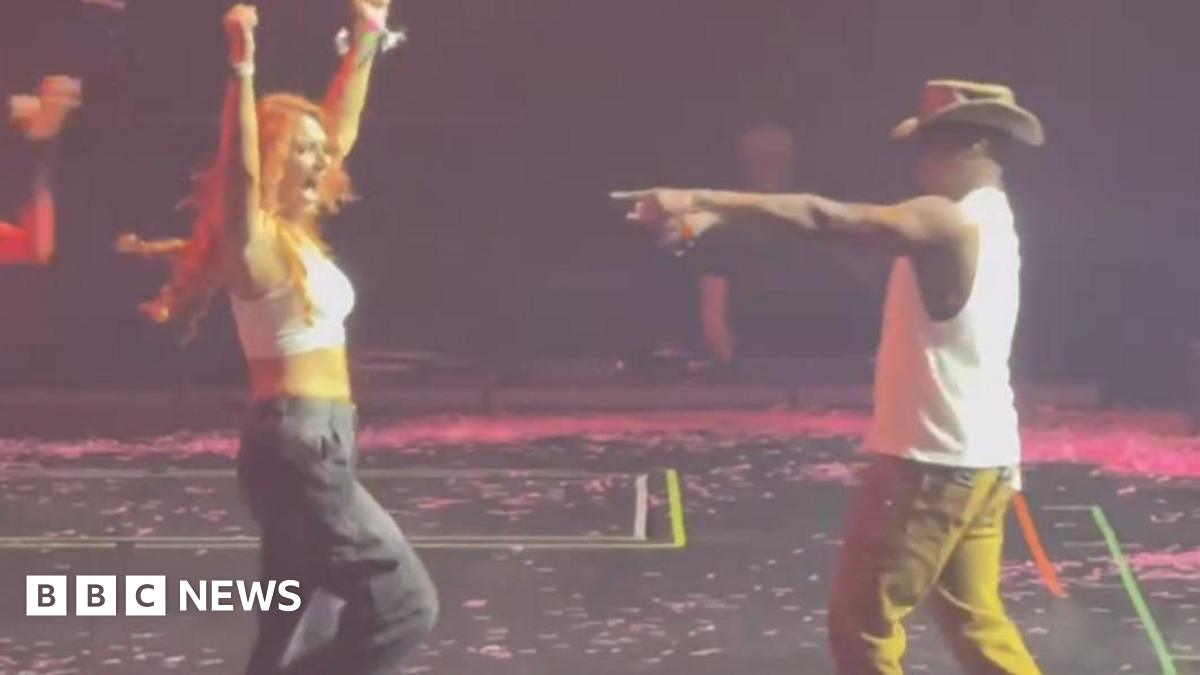 A woman with long red hair, a white tank top and black cargo trousers stands on a black stage stage with her arms in the air. To the right singer Ne-Yo, wearing a white vest top, mustard coloured trousers and a brown cowboy-style hat points towards her with his right hand.