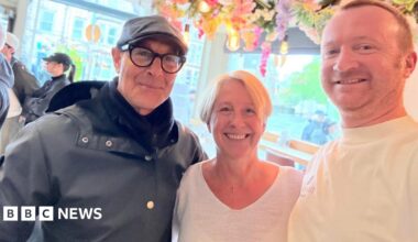 Stanley Tucci, wearing a cap and glasses, with a woman with short blonde hair and a man with short auburn hair, both wearing white T-shirts and standing in a cafe.