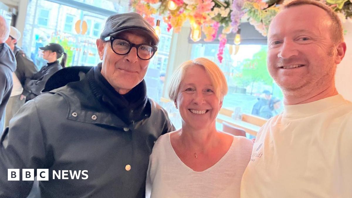Stanley Tucci, wearing a cap and glasses, with a woman with short blonde hair and a man with short auburn hair, both wearing white T-shirts and standing in a cafe.
