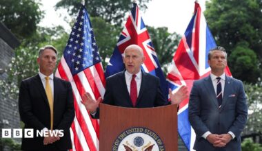 John Healey speaks and gestures with his hands at a podium at the US embassy, flanked by Richard Marles and Pete Hegseth. The US, UK and Australia flags are posted behind them.