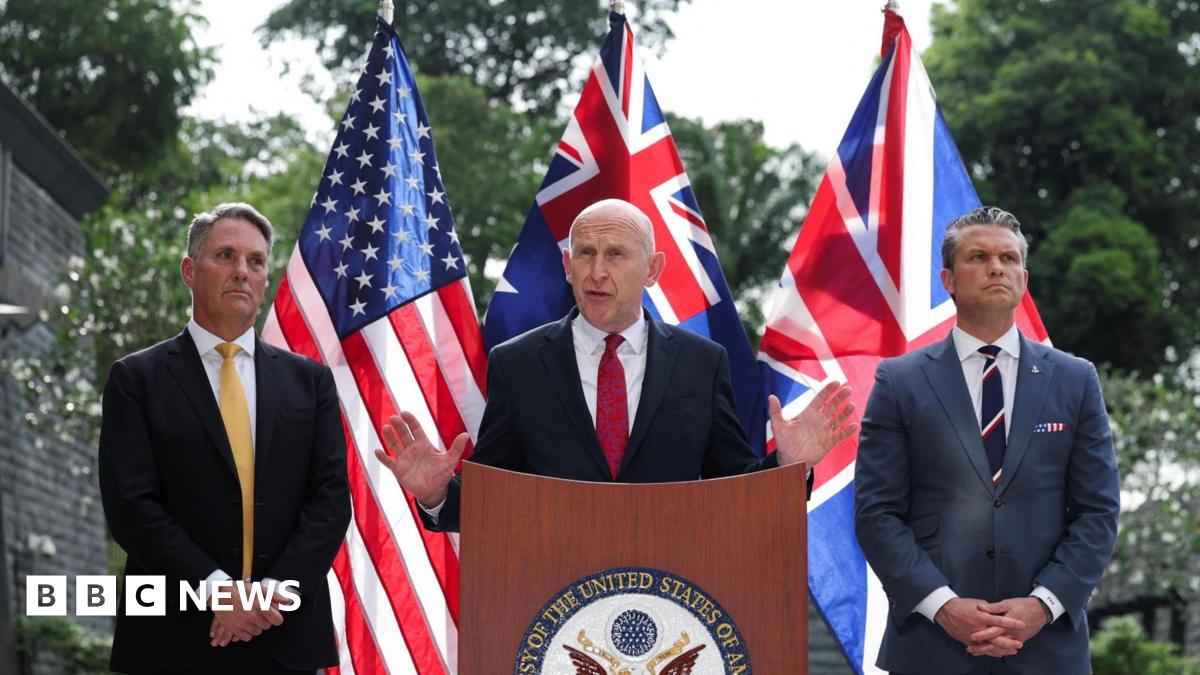 John Healey speaks and gestures with his hands at a podium at the US embassy, flanked by Richard Marles and Pete Hegseth. The US, UK and Australia flags are posted behind them.