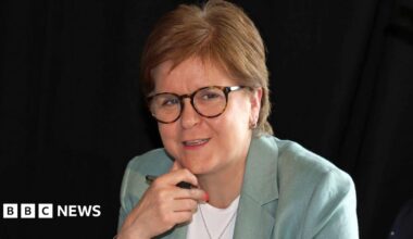 Nicola Sturgeon sitting down, wearing a pale green suit over a white top. She has her hand underneath her chin and is holding a pen.