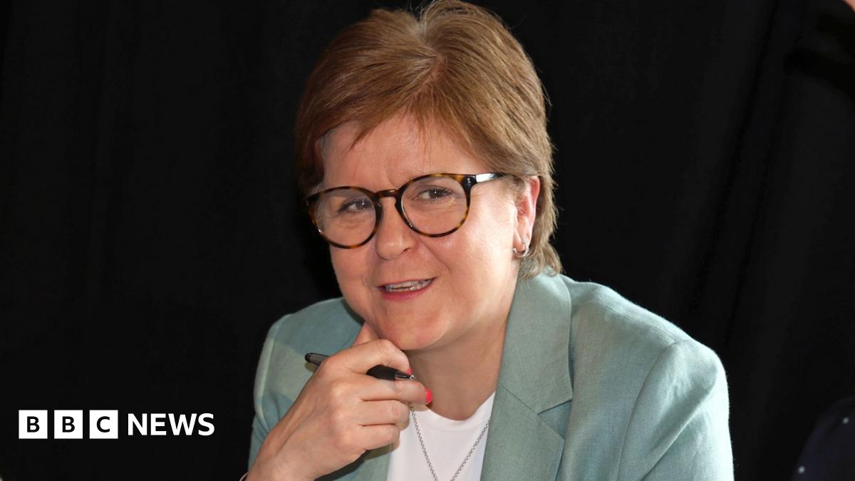 Nicola Sturgeon sitting down, wearing a pale green suit over a white top. She has her hand underneath her chin and is holding a pen.