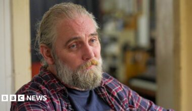 Adam, a man with long hair and a beard, wearing a chequered shirt, looks at the camera with a neutral expression