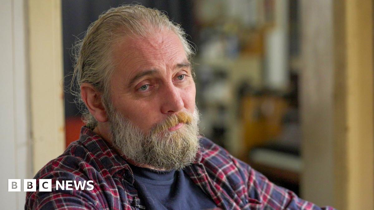 Adam, a man with long hair and a beard, wearing a chequered shirt, looks at the camera with a neutral expression