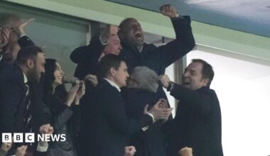 Prince William pictured with his arms in the air celebrating with people around him in the football stands also clapping, smiling and hugging.