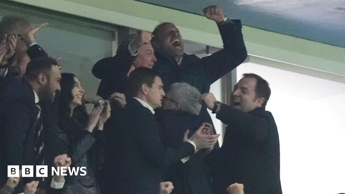 Prince William pictured with his arms in the air celebrating with people around him in the football stands also clapping, smiling and hugging.