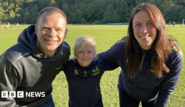 Edie, nine, stands smiling between her dad Matt and her coach Amy Sherratt on the grass at Oakamoor Cricket Club. She wears a navy Oakamoor Cricket Club training top with the club's oak leaf badge. Behind them, junior players take part in a training session against a backdrop of wooded hillside on a bright, sunny evening.