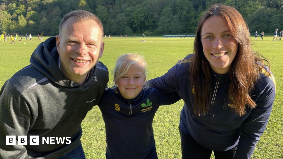 Edie, nine, stands smiling between her dad Matt and her coach Amy Sherratt on the grass at Oakamoor Cricket Club. She wears a navy Oakamoor Cricket Club training top with the club's oak leaf badge. Behind them, junior players take part in a training session against a backdrop of wooded hillside on a bright, sunny evening.