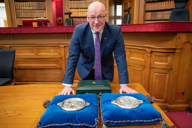 John Swinney in blue suit, white shirt and purple tie, standing over two versions of the Great Seal of Scotland.