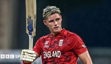 Jacob Bethell, in red team shirt, raises his bat after scoring a century for England