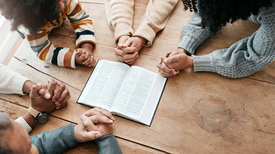 A multigenerational group prays around an open Bible at a wooden table