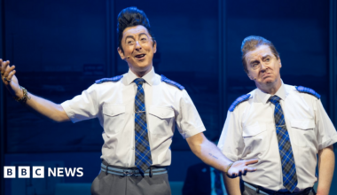 Two performers in matching airline-style uniforms perform on stage in The High Life: The Musical, gesturing as they sing under blue stage lighting.