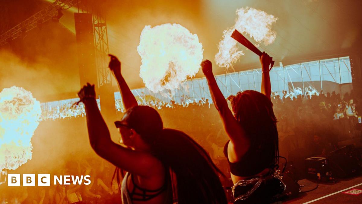 Two women on stage in a marquee raise their arms as flames shoot up on the first day of Love Saves The Day. A large crowd inside the marquee can be seen illuminated in the flames