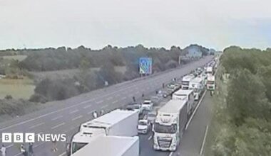 A motorway camera view showing a queue on one carriageway, People can be seen out of their cars.